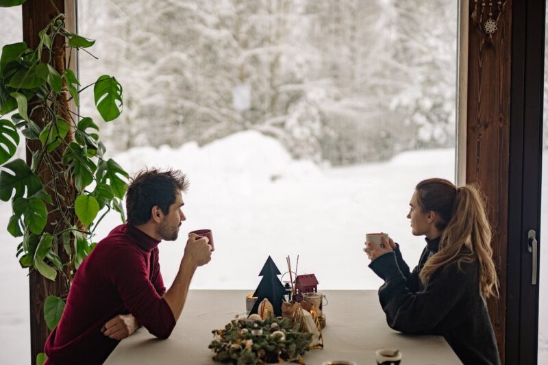 PH-EN-Website-Passivhaus-Snow-Outside A couple in front of a window in their dining room enjoying warm cups of coffee looking out to the snowy landscape.